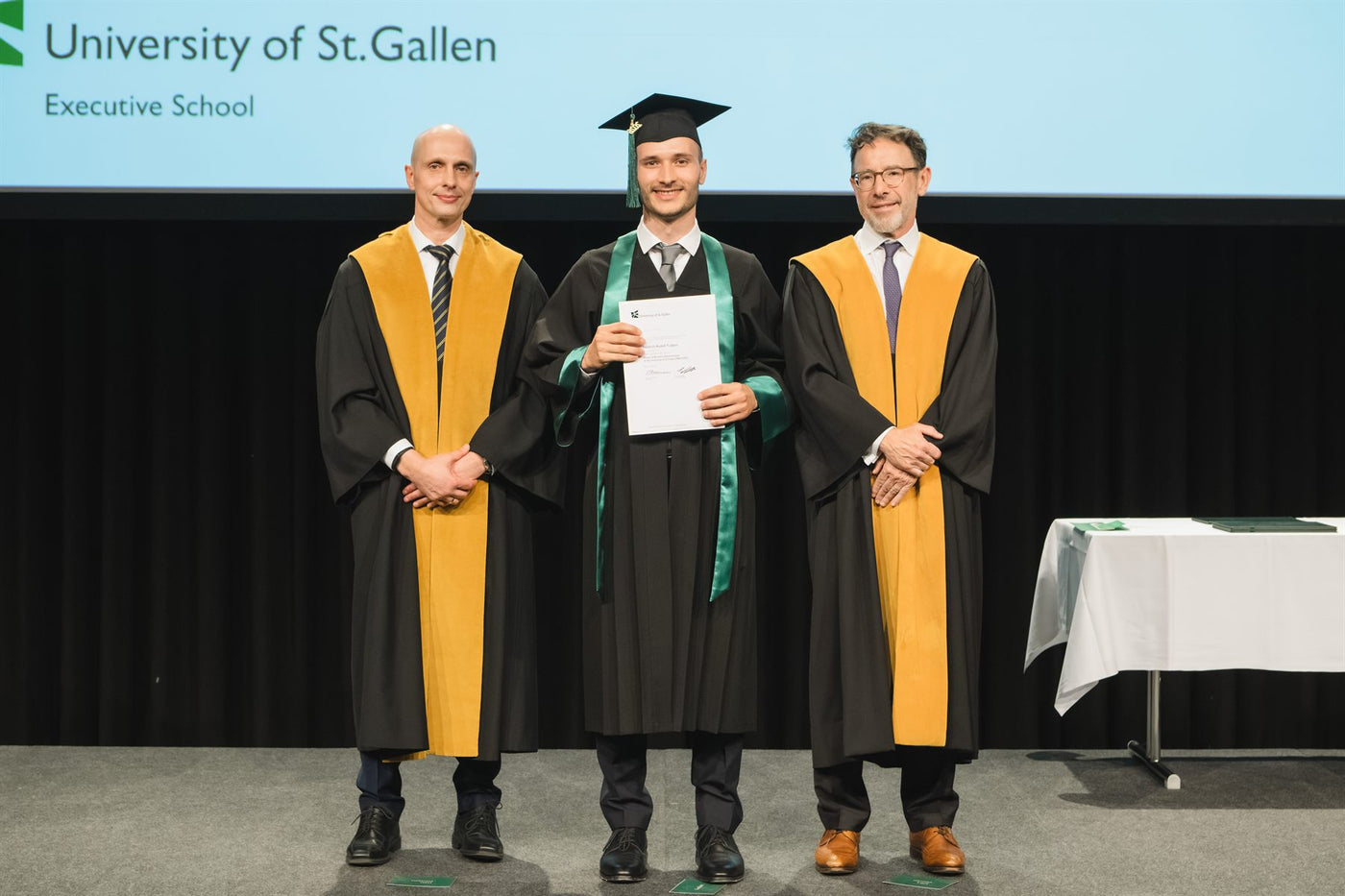 Friedrich Trabert in Talar und Barett mit seiner Urkunde, flankiert von zwei Vertretern der Executive School, vor der Leinwand der Universität St. Gallen.